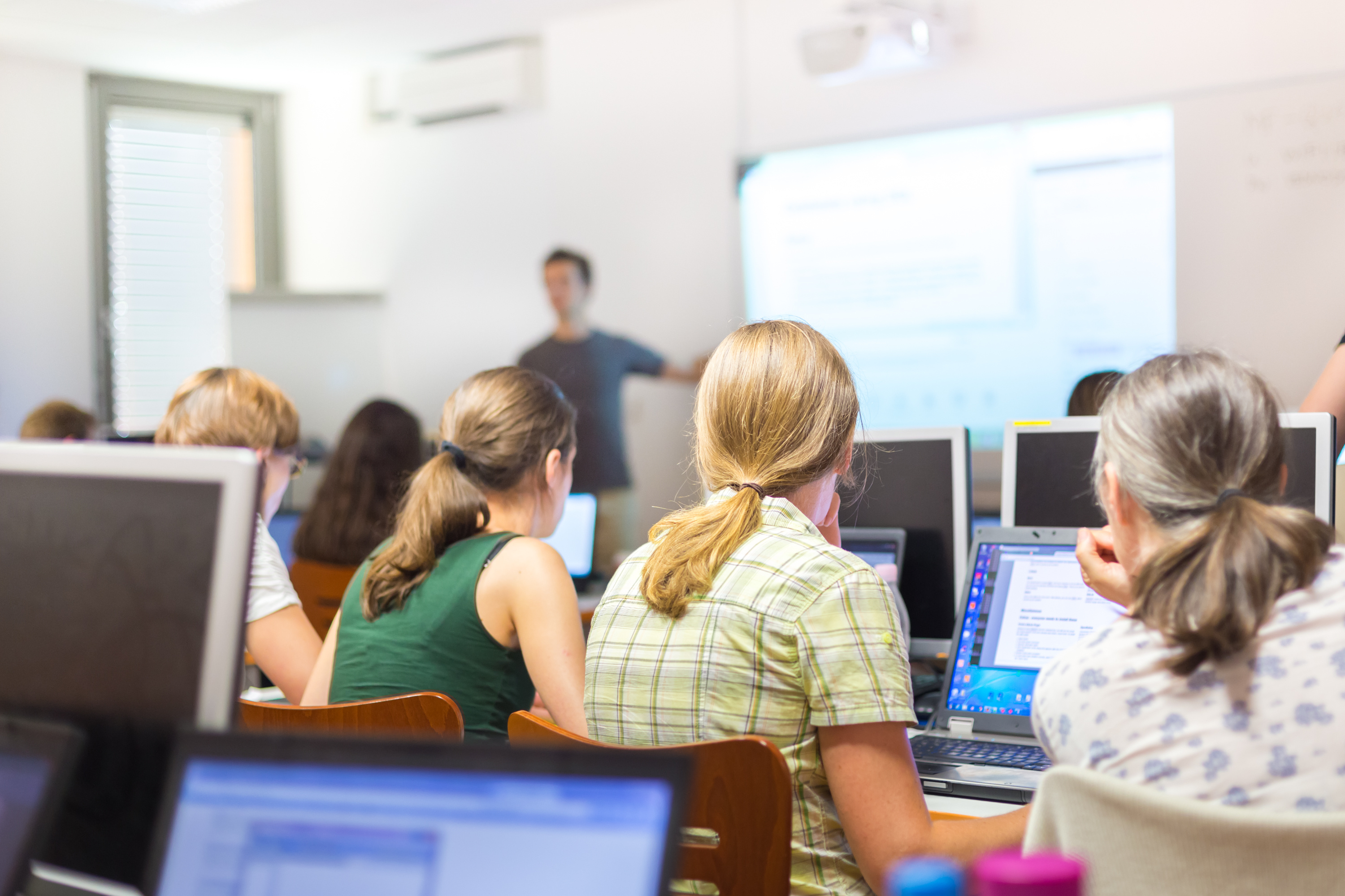 Workshop at university. Rear view of students sitting and listening in lecture hall doing practical exercises on their laptop computers. Tutor explaining tasks on white board.