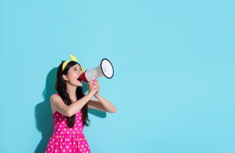 happy beautiful woman using megaphone speaking information and wearing pink cute dress in blue wall background.